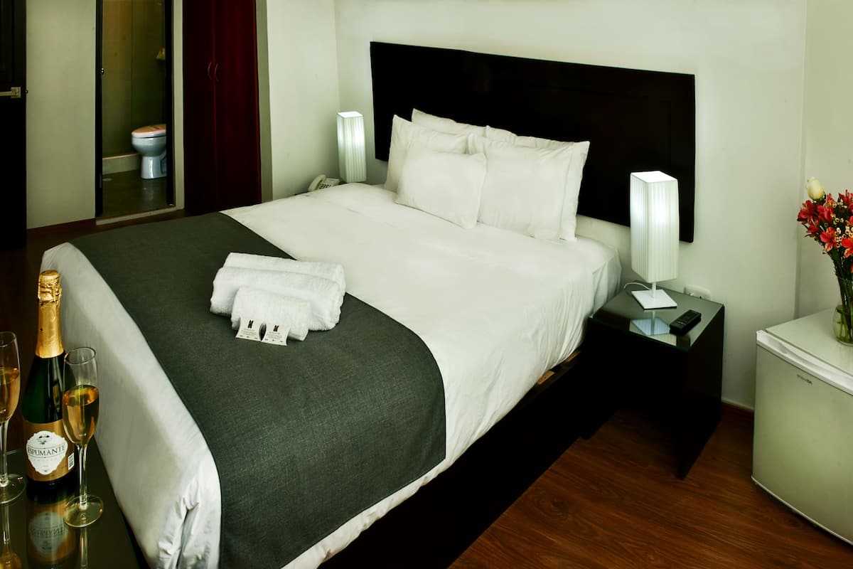 A hotel room with a large bed, a nightstand with a lamp, a small refrigerator, and a vase with flowers.