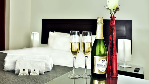 A neatly made bed with white linens, a bottle of champagne, two glasses of champagne, and a red vase with a white flower on a hotel room table. - Riviera Hoteles Ejecutivo (Arequipa)