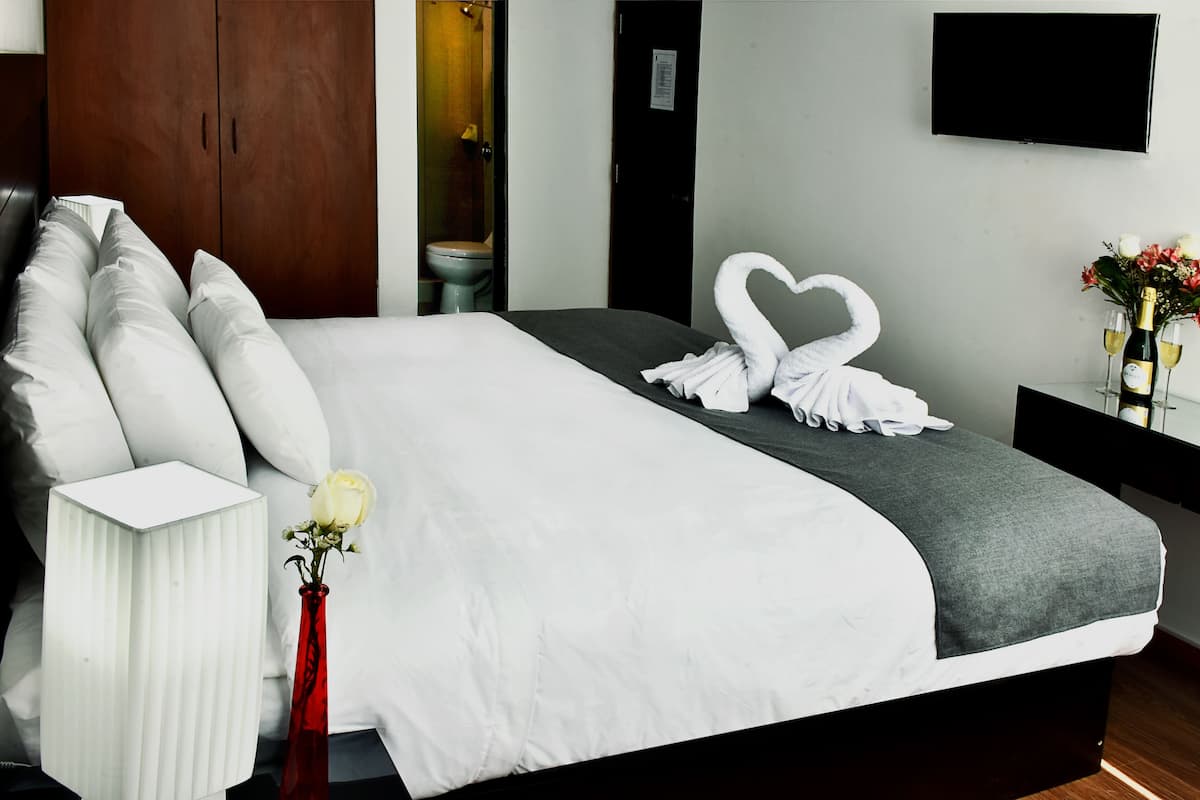 A neatly made bed with white and grey bedding, a red vase with a single white rose, and a heart-shaped towel arrangement on the bed.