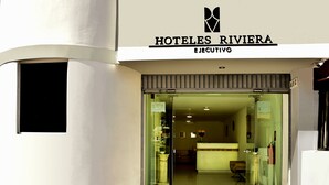The entrance to Hotel Riviera Executive with a glass door, a reception desk, and a visible interior with a sofa and a vase of flowers. - Riviera Hoteles Ejecutivo (Arequipa)