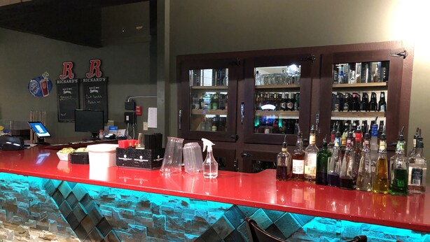 Una zona de bar con mostrador rojo, pared de piedra y varias botellas.