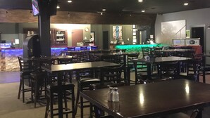 Un restaurante con varias mesas y sillas, una zona de bar con mostrador y taburetes, y un televisor instalado en un pilar.
