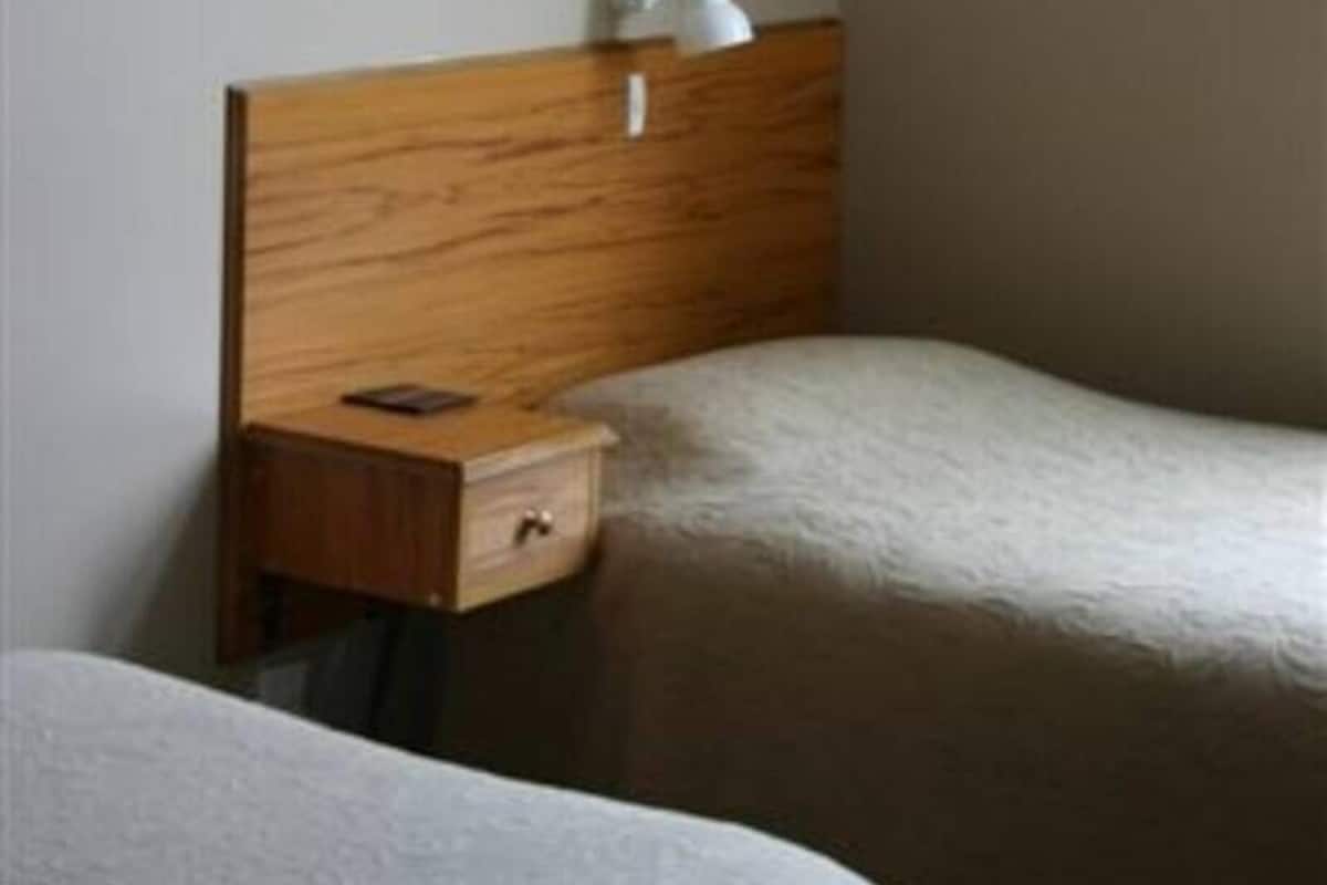 A hotel room with two single beds, a wooden headboard, a bedside table with a lamp, and a small drawer.