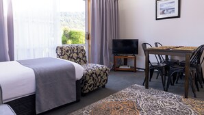 A room with a bed, a TV, a dining table with chairs, and a view of greenery outside. - Apostles View Motel (Greymouth)