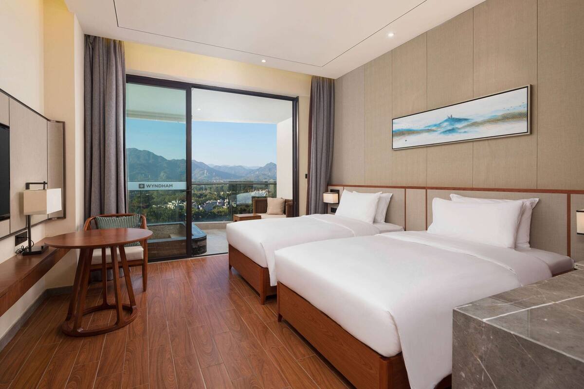 A hotel room with two beds, a wooden table, a chair, a large window with a view of mountains, and a painting on the wall.