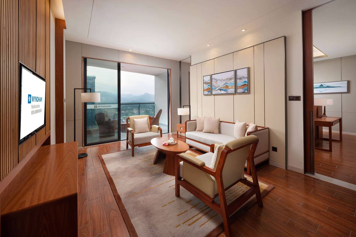 A modern hotel room with a wooden reception desk, a sofa, two armchairs, a coffee table, and a large window offering a city view.