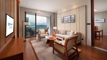A modern hotel room with a wooden reception desk, a sofa, two armchairs, a coffee table, and a large window offering a city view.