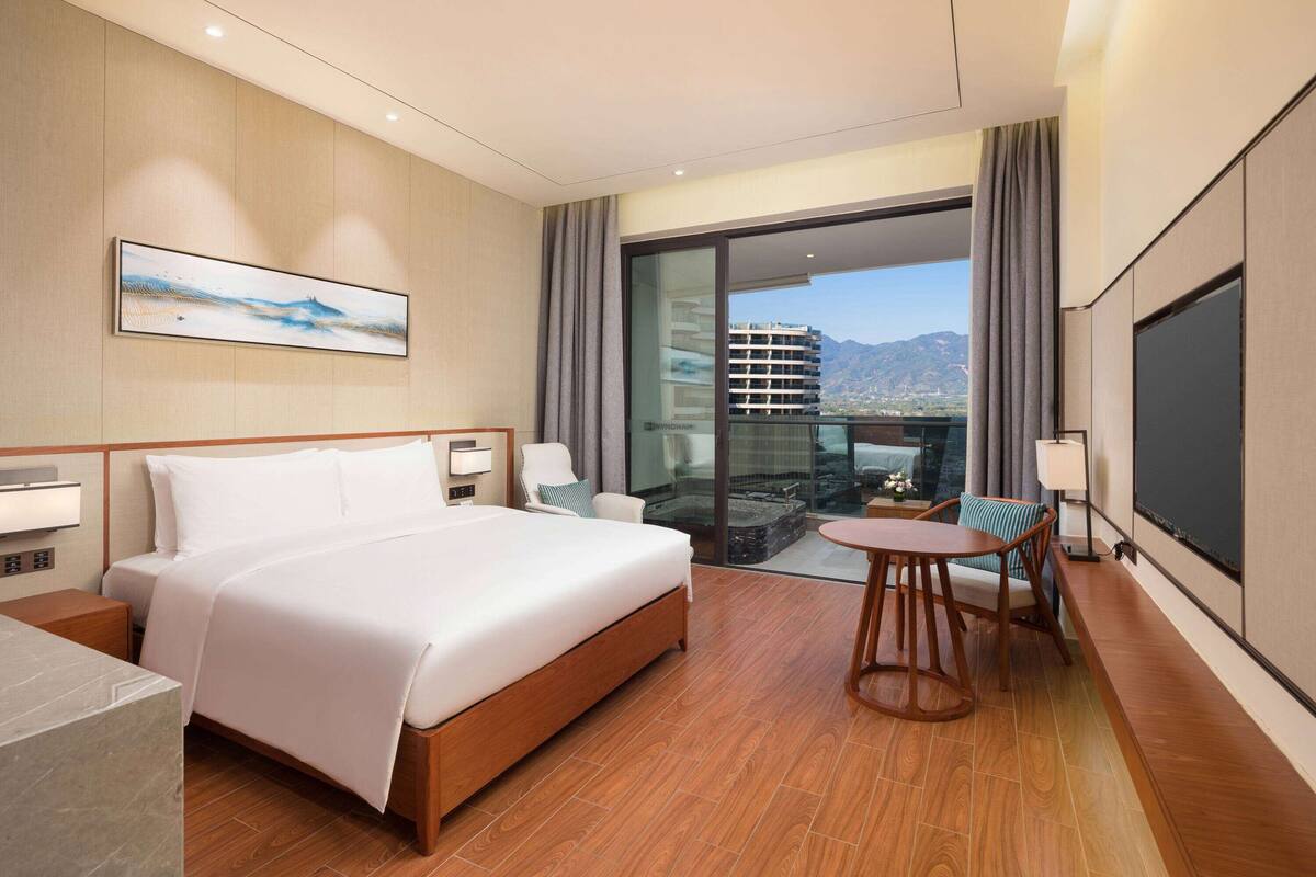 A hotel room with a large bed, a small table with chairs, a TV, and a balcony with a city view.