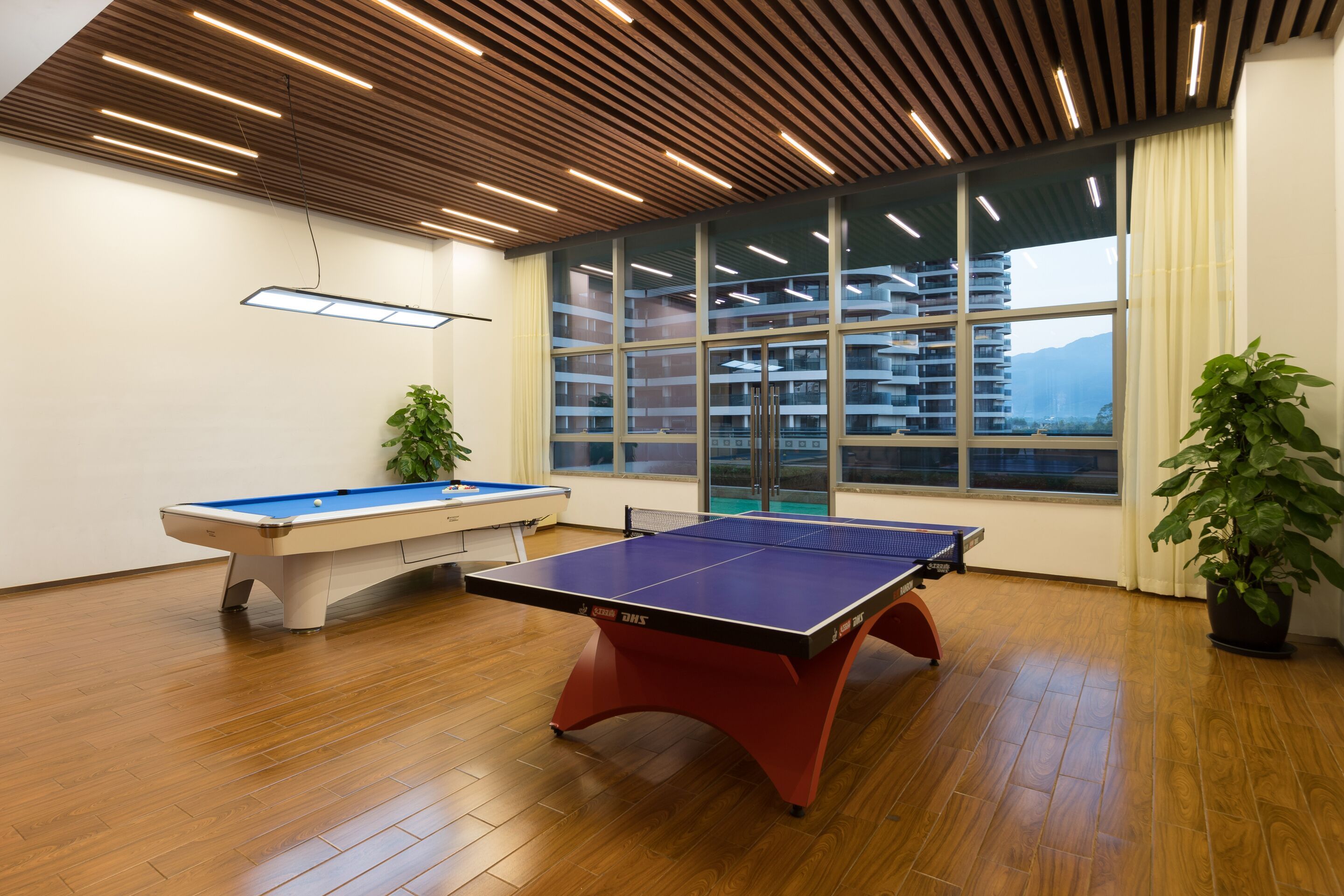 A room with a ping pong table, a pool table, and a large window with a view of buildings.