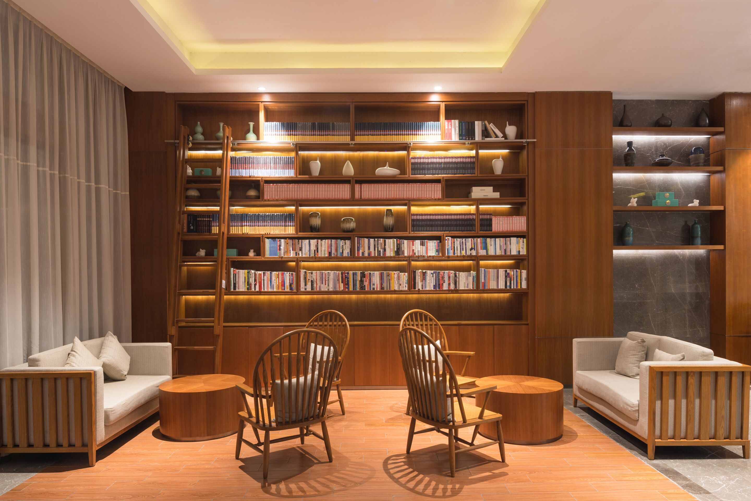 A modern library with wooden bookshelves, two sofas, and wooden chairs.