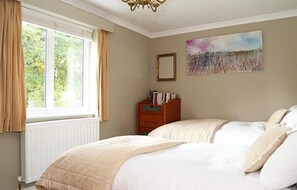 Une chambre avec un grand lit, une table de chevet avec des livres, une fenêtre avec des rideaux et un tableau au mur.