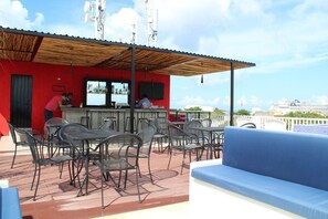Een terras met metalen stoelen en een blauwe bank, een rood gebouw met een zwarte deur, en een uitzicht op een cruiseschip op de achtergrond.