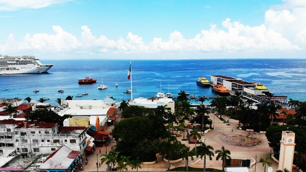 A coastal cityscape with a cruise ship docked, a marina with various boats, and a central plaza with palm trees and a clock tower. - Hotel Pal Cozumel (Cozumel)