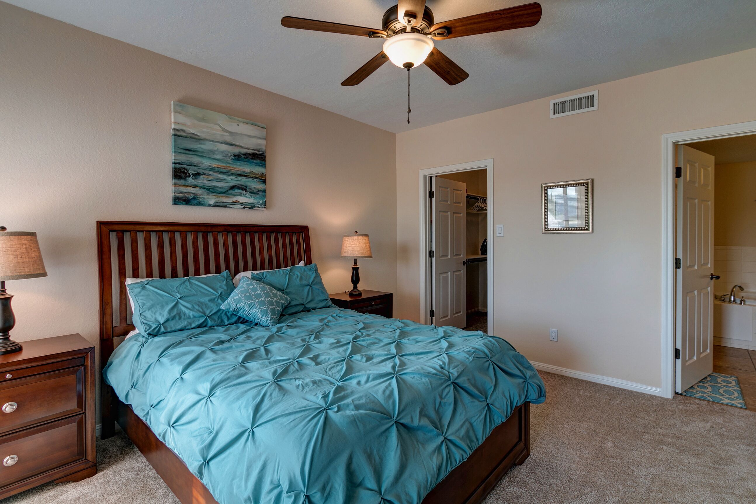 A bedroom with a bed, a ceiling fan, a nightstand, a lamp, a painting, and an open closet door.