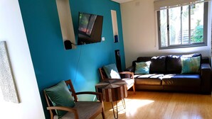 A living room with a blue wall, a TV mounted on it, a sofa with cushions, a wooden chair, and a small wooden table. - Beautiful Beach-side cottage with lap pool in North Maroubra close to Coogee (Maroubra)