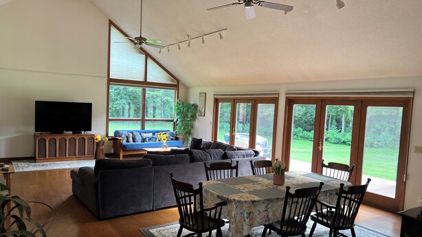 A spacious living room with a large TV, a sofa set, a dining table with chairs, and a ceiling fan. - Spacious Home nestled along the Blackberry River in Northwest CT (East Canaan)