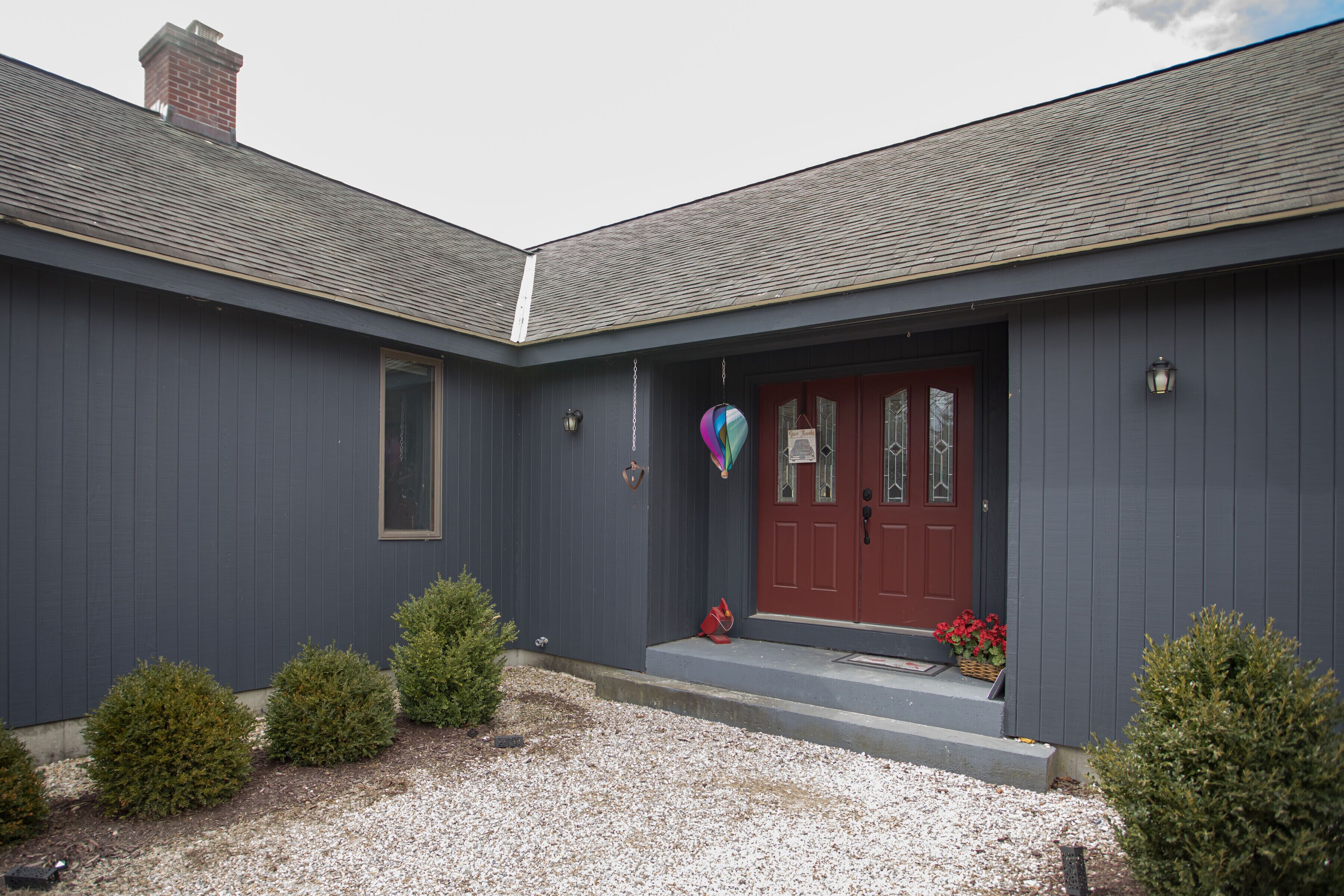 Une maison à deux étages, à l’extérieur de couleur gris foncé, avec une porte d’entrée rouge et un petit porche agrémenté d’un panier rempli de fleurs rouges.