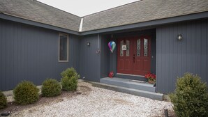 A two-story house with a dark gray exterior, a red front door, and a small porch area with a basket of red flowers. - Spacious Home nestled along the Blackberry River in Northwest CT (East Canaan)