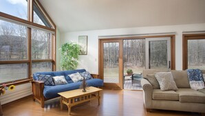 A spacious living room with a blue sofa, a beige sofa, and a wooden coffee table. There are large windows and a sliding glass door leading to a deck. - Spacious Home nestled along the Blackberry River in Northwest CT (East Canaan)