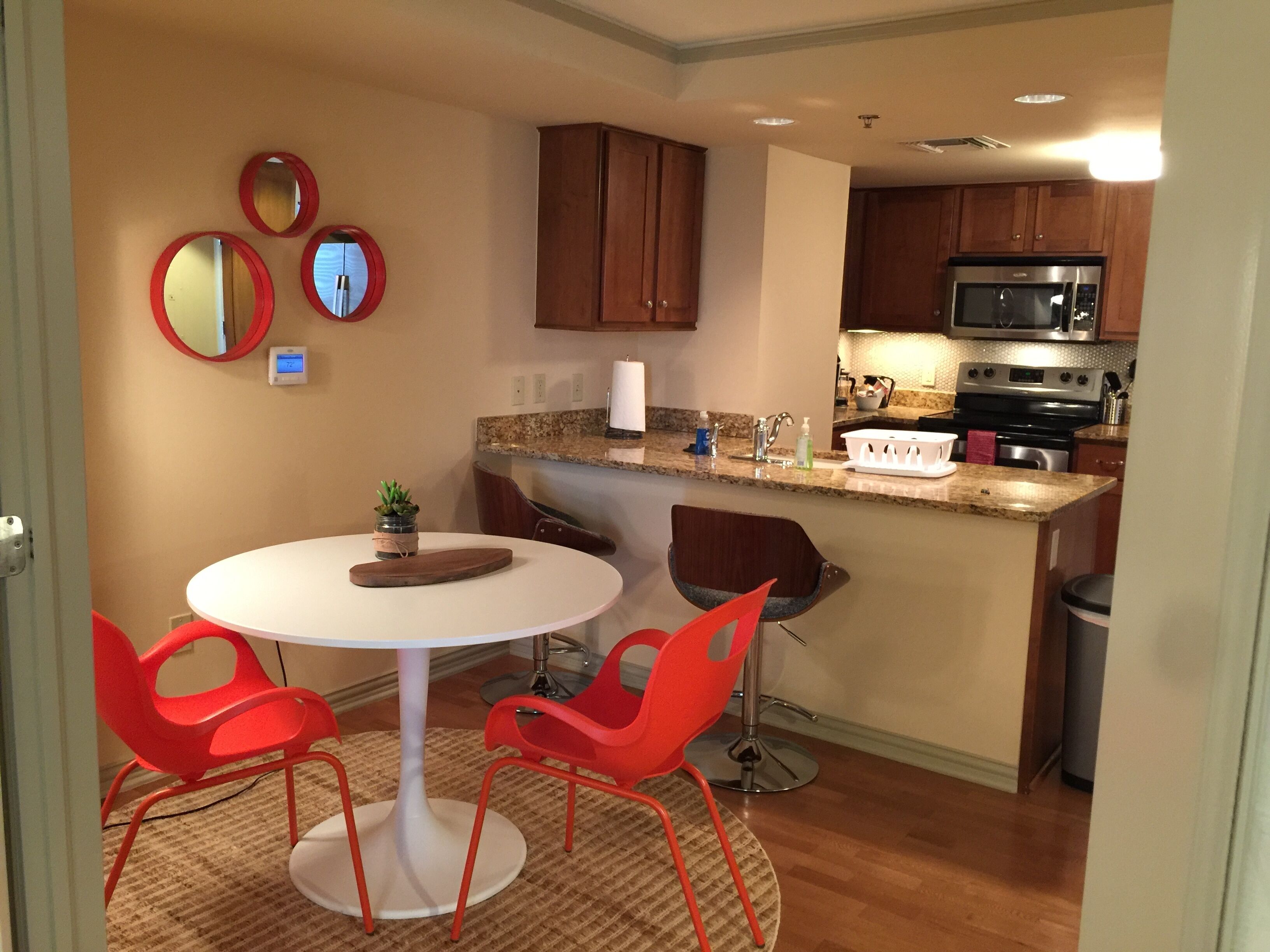 A compact kitchen with a microwave, stove, and sink. There is a small dining area with a white table and red chairs.