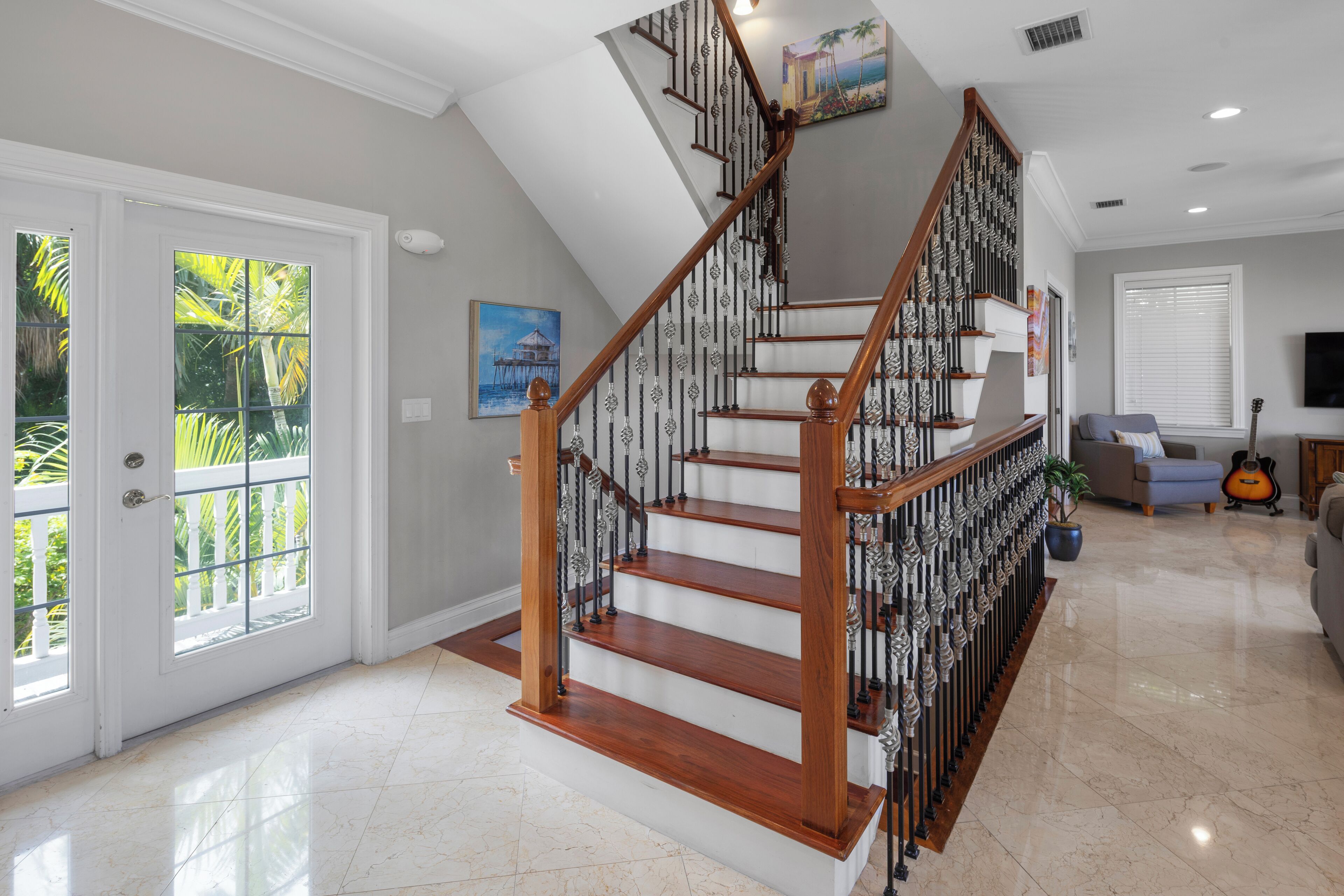 A spacious living area with a wooden staircase, a large window with blinds, a guitar, and a painting on the wall.