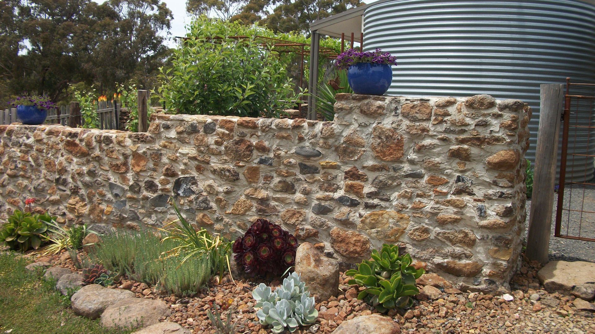 Un mur de pierre bordé d’un jardin où l’on trouve différentes espèces de plantes et un élément décoratif aquatique.