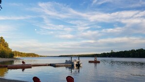 A lakeside scene with two red chairs on the shore, a dock with a boat, and a calm water body under a partly cloudy sky. - Perfect Lakefront Getaway ~ Beautiful Private Setting!  (Dedham)