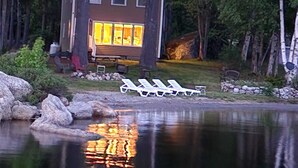 A cabin with a large window, a deck with lounge chairs, and a reflection in the water. - Perfect Lakefront Getaway ~ Beautiful Private Setting!  (Dedham)