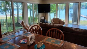 A spacious living room with a large window overlooking a lake, a television, and a dining area with a table set for two. - Perfect Lakefront Getaway ~ Beautiful Private Setting!  (Dedham)