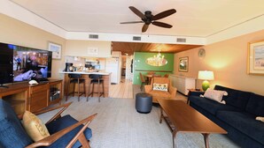 A living room with a TV, sofa, chairs, a coffee table, a ceiling fan, and a kitchen area in the background. - Traditional 1 Bedroom Oceanfront Condo with Free WiFi, an Outdoor Pool, and a Wonderful View Located in Midtown and Mere Steps to the Beach! (Ocean City)