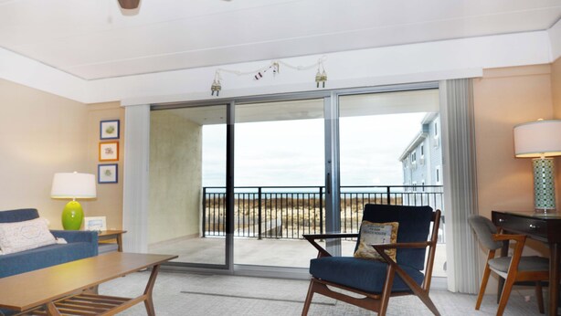 A room with a sofa, a chair, a table, a lamp, and a view of the beach through a sliding door. - Traditional 1 Bedroom Oceanfront Condo with Free WiFi, an Outdoor Pool, and a Wonderful View Located in Midtown and Mere Steps to the Beach! (Ocean City)