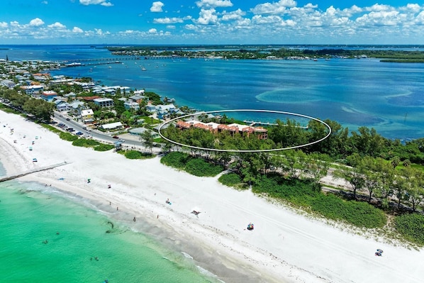 An aerial view of a coastal area with a beach, a marina, and a cluster of buildings with a circular boundary.