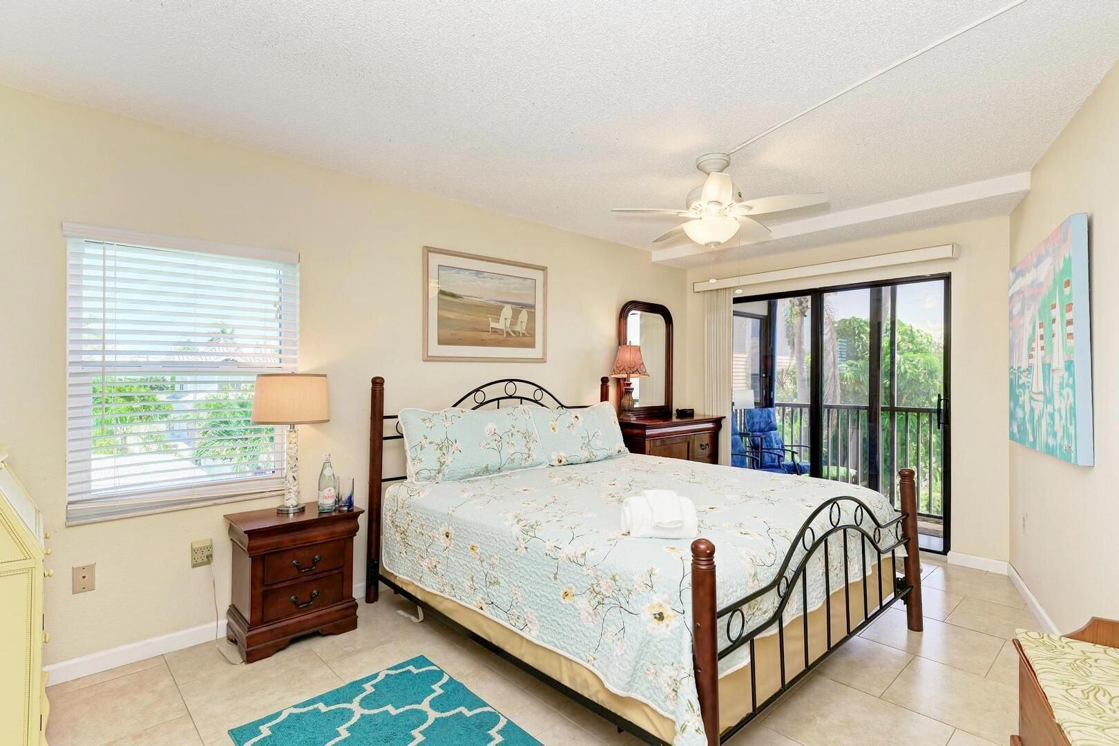A bedroom with a bed, a nightstand, a dresser, a window with blinds, a ceiling fan, and a painting on the wall.