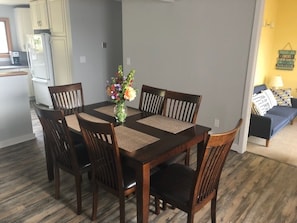 A dining area with a wooden table and chairs, a vase with flowers, and a kitchen in the background.