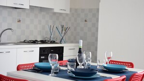 Cuina moderna amb armaris blancs, fornet a gas i rentavaixelles. Hi ha un menjador amb plats blaus i cadires vermelles.