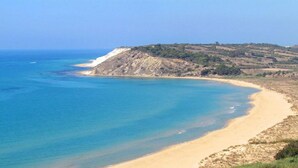 Paisatge costaner amb una platja sorrenca, aigües blaves cristal·lines i un relleu amb zones de muntanya al fons.