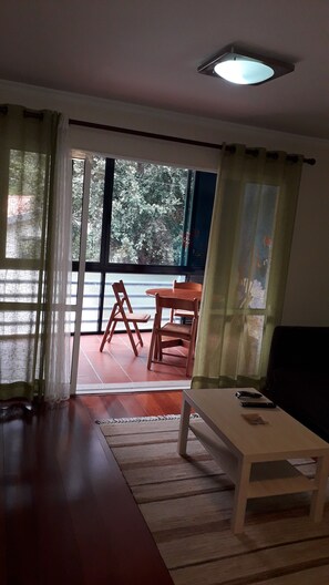 A living room with a sofa, a coffee table, and a dining table with chairs. There is a sliding glass door leading to an outdoor area with a view of trees.