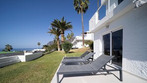 Un'area lounge esterna moderna con tre lettini, un edificio bianco con ampie finestre e palme.