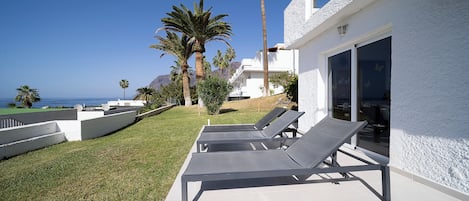A modern outdoor lounge area with three sunbeds, a white building with large windows, and palm trees.
