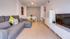 A modern living room with a grey sectional sofa, a dining table with chairs, and a flat-screen TV on a white cabinet. - Apt. with 4 pools, very close to Fenals beach, 6 pax, air conditioning and free wifi. (Lloret de Mar)