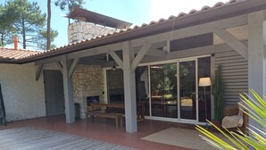Ein überdachter Patio-Bereich mit Kamin aus Stein, Holzmöbeln und einer großen Schiebeglastür, die zu einem Außenbereich führt.