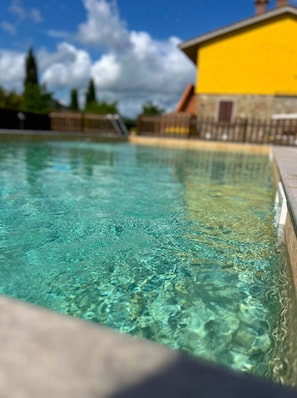 A clear swimming pool with a stone edge and a yellow building in the background.