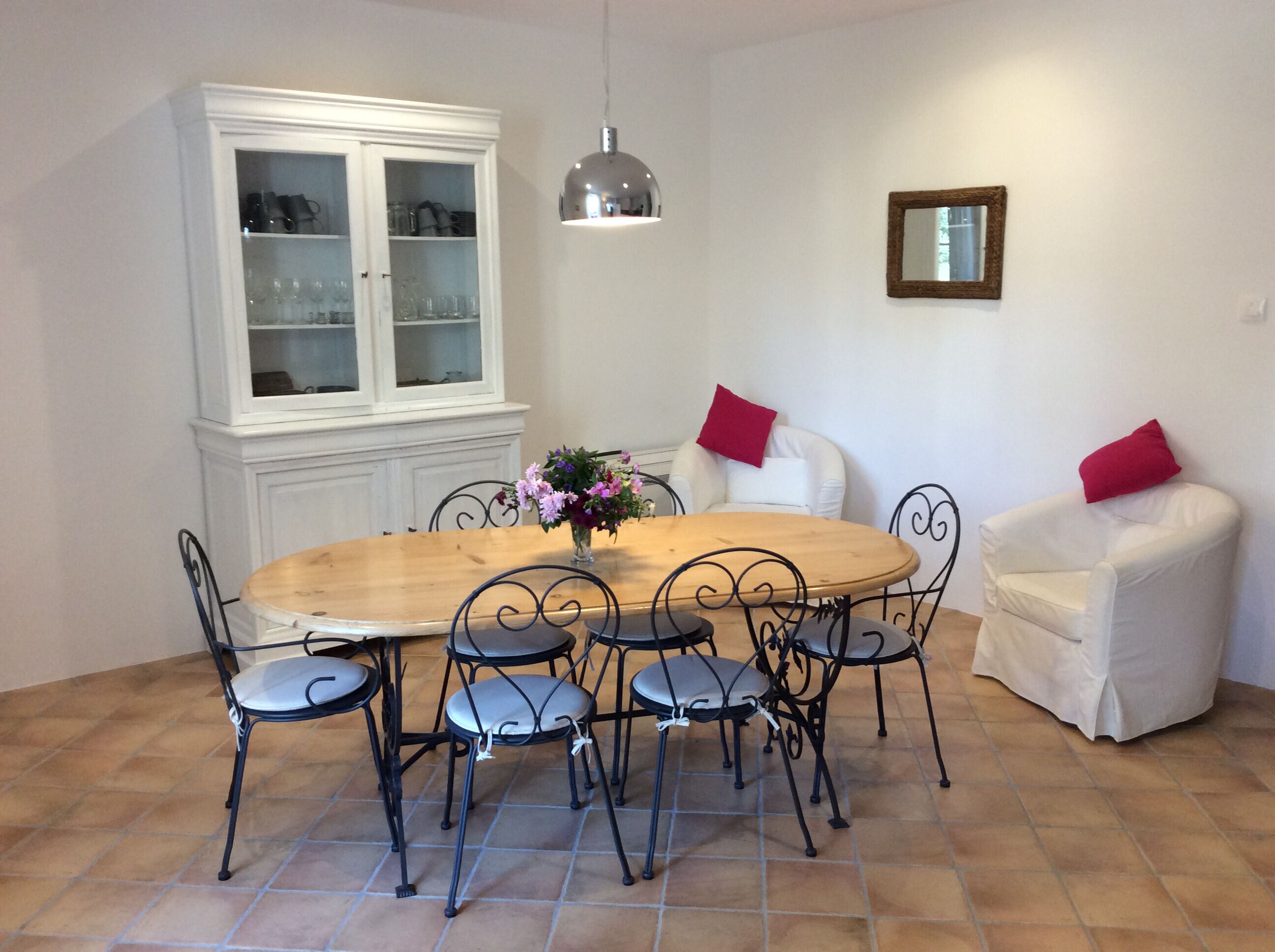 A dining area with a round wooden table surrounded by four chairs with metal frames and cushioned seats. There is a white cabinet with glass doors to the left, and a white armchair with red cushions to the right. A pendant light hangs above the table.