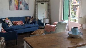 Ein Wohnzimmer mit einem blauen Sofa, einem Holztisch, einem Sessel mit einer rosa Decke, einem großen Spiegel und einem Fenster mit Blick auf Gebäude.