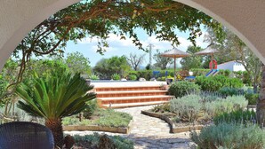 Un sentier de jardin mène à une zone de piscine agrémentée de marches, le tout étant entouré de diverses plantes et d'un fauteuil en osier.