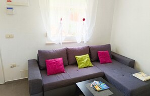 Un sofá gris con tres cojines, una ventana con cortinas translúcidas, una mesita con revistas y un termostato fijado a la pared.