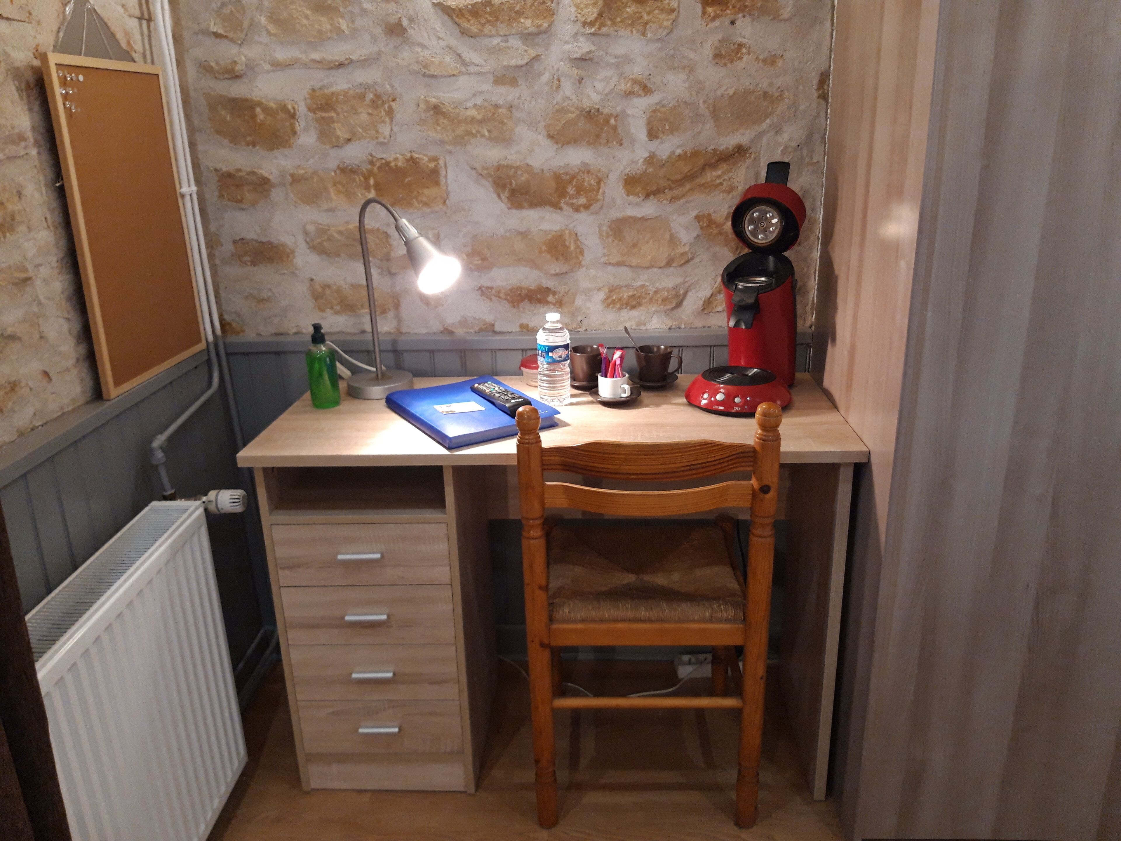Una habitación pequeña y rústica con un escritorio de madera, una silla, un radiador, un tablón de corcho, una lámpara, una cafetera roja y una botella de agua.