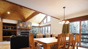 A dining area with a wooden table and chairs, a stone fireplace, and large windows offering a view of snow-covered trees. - 61 Jabberwocky Private vacation home 5 bedroom, pet friendly clubhouse amenities (Fraser)