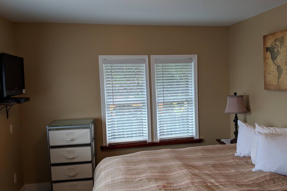 A bedroom with a bed, a dresser, a lamp, a map on the wall, and a window with blinds.