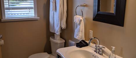 Une salle de bain avec des toilettes, un lavabo et un miroir. Des serviettes sont suspendues sur un porte-serviettes, et il y a une fenêtre avec des stores.